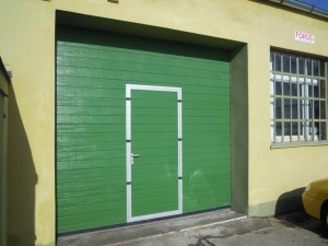 Brama przemysłowa segmentowa Wiśniowski Makropro z drzwiami przejściowymi, struktura panela: woodgrain, kolor: RAL:6005